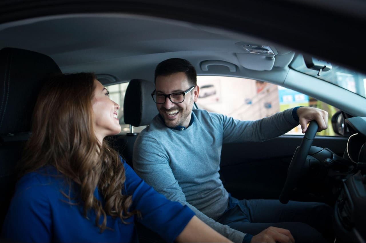 Casal de uma mulher e um homem se olhando, ambos vestindo suéter azuis e sorridentes. O homem está no banco de motorista e a mulher no banco de passageiro de um veículo próprio.