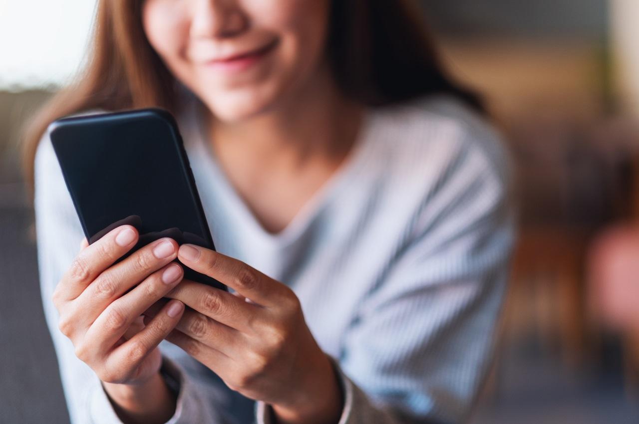 Mulher sorridente segurando um celular com as duas mãos. Ela veste uma blusa de manga cumpridas branca.