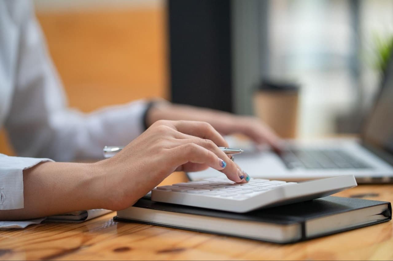 Pessoa (seu rosto não aparece na imagem) calcula adiantamento salarial em calculadora e anota valores no laptop aberto na mesa de trabalho.