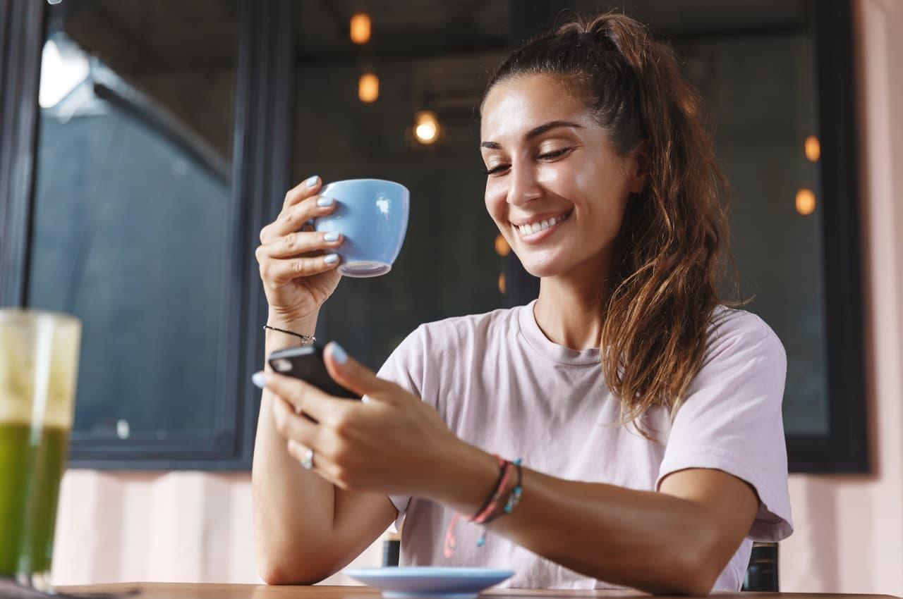 Mulher sorridente segurando uma xícara azul em uma das mãos e um celular em outra. Ela está lendo sobre amortização de empréstimo.