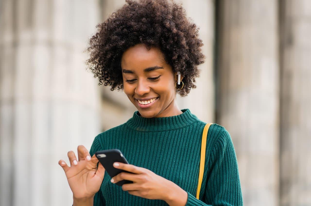 Mulher sorridente está em pé na calçada de uma rua, digitando no celular. Ela veste blusa de gola alta verde e fones de ouvido sem fio.