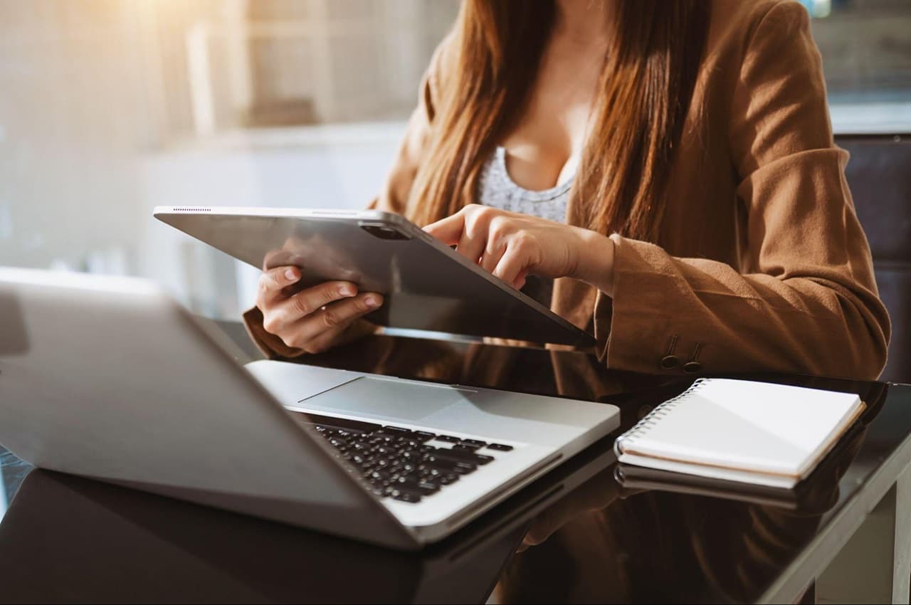 Mulher vestindo casaco de sarja marrom, utilizando tablet para cancelar empréstimo consignado. Ela está no escritório do trabalho com um computador e caderno de anotações aberto em cima da sua mesa.