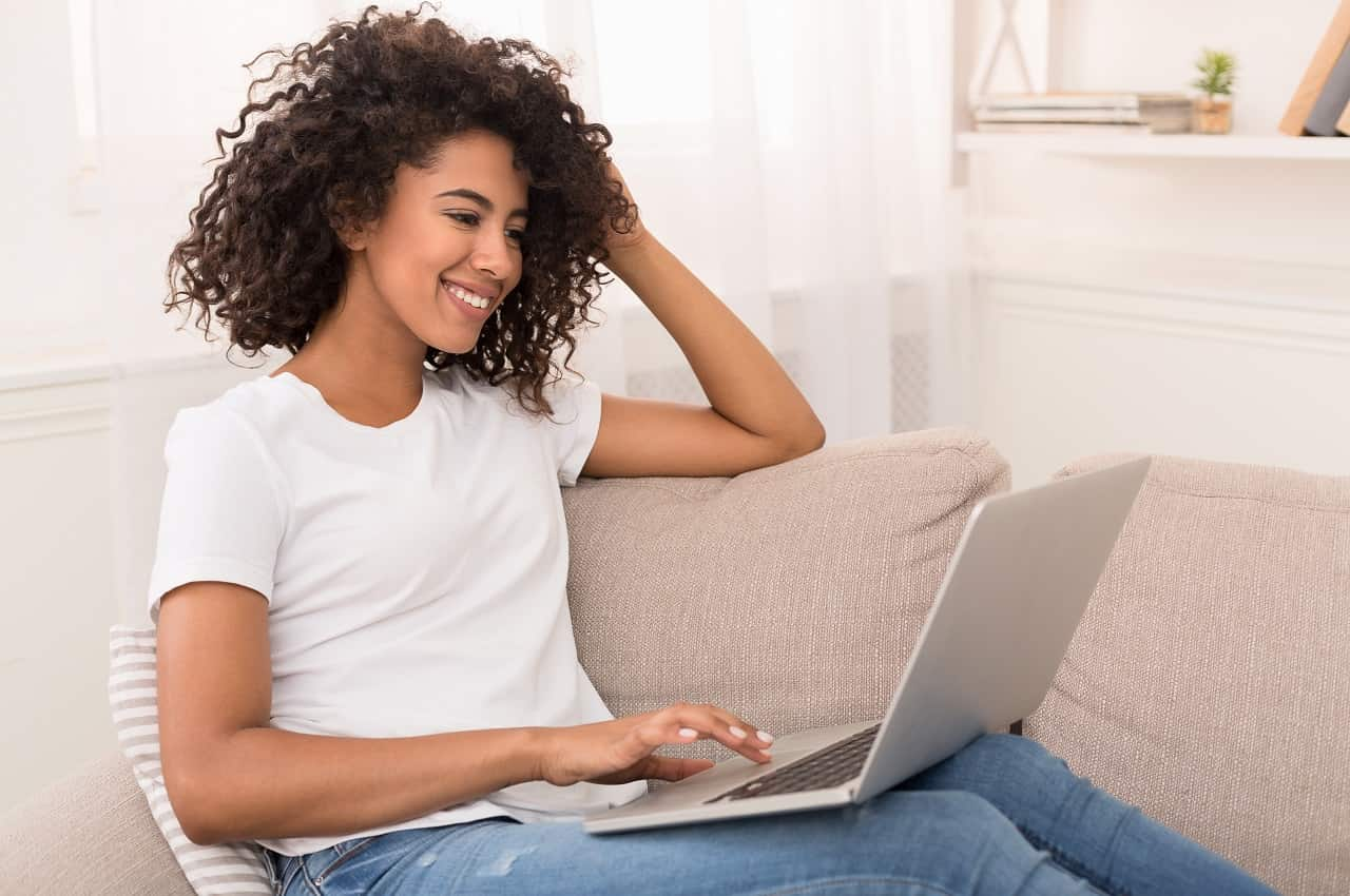 Mulher sorridente vestindo blusa básica branca, calça jeans e sentada no sofá, enquanto utiliza um laptop no seu colo.