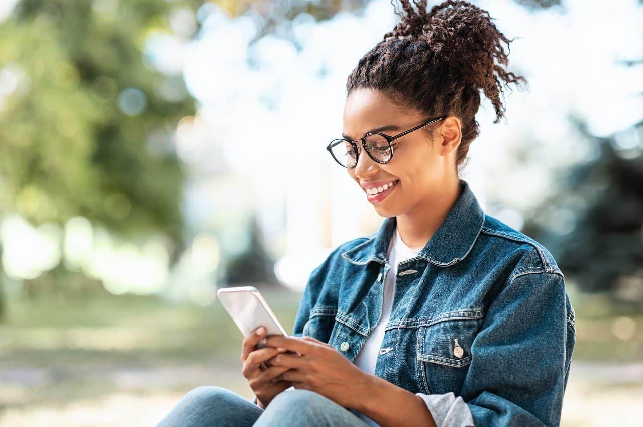 Mulher sorridente sentada em um parque e pesquisando no celular como funciona empréstimo consignado privado. Ela veste jaqueta e calça jeans, com uma blusa básica branca e óculos de grau.