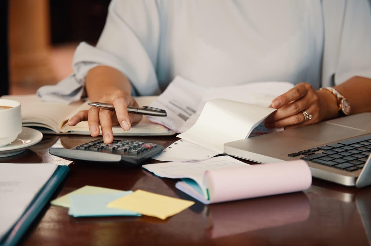 Pessoa (seu rosto não aparece na imagem) usa calculadora e um laptop para calcular como liberar margem consignável. Em uma mesa tem post-it, documentos e extratos bancários.