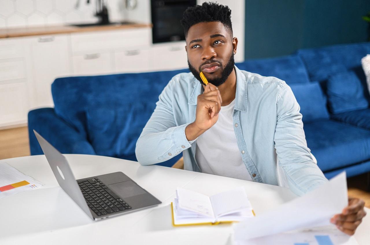 Homem negro de cabelos curtos pretos com expressão concentrada, apoiando um lápis no queixo e olhando para o desconto do empréstimo consignado na rescisão. Ele veste blusa básica branca, jaqueta jeans aberta e está na mesa da sala de jantar.