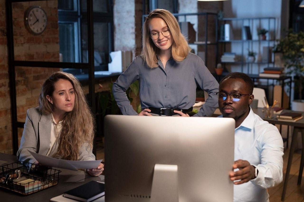 Grupo de três pessoas sorridentes (duas mulheres e um homem) olhando para o monitor de um computador. As mulheres seguram folhas de papel, enquanto o homem ajusta o monitor.