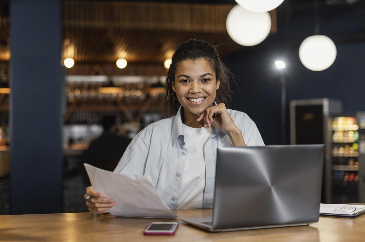 Mulher sorridente de cabelos presos em coque, camisa social branca e sentada no escritório. Ela segura papel de empréstimo para CLT e usa um laptop aberto. 