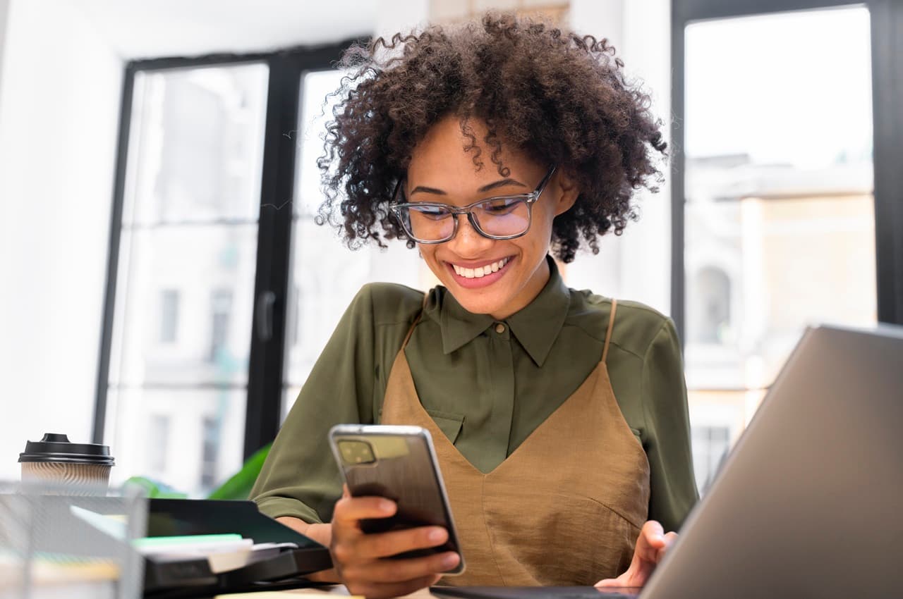 Mulher sorridente de óculos de grau, digitando no celular e no computador do escritório. Ela veste camisa verde musgo social e está no ambiente de trabalho.