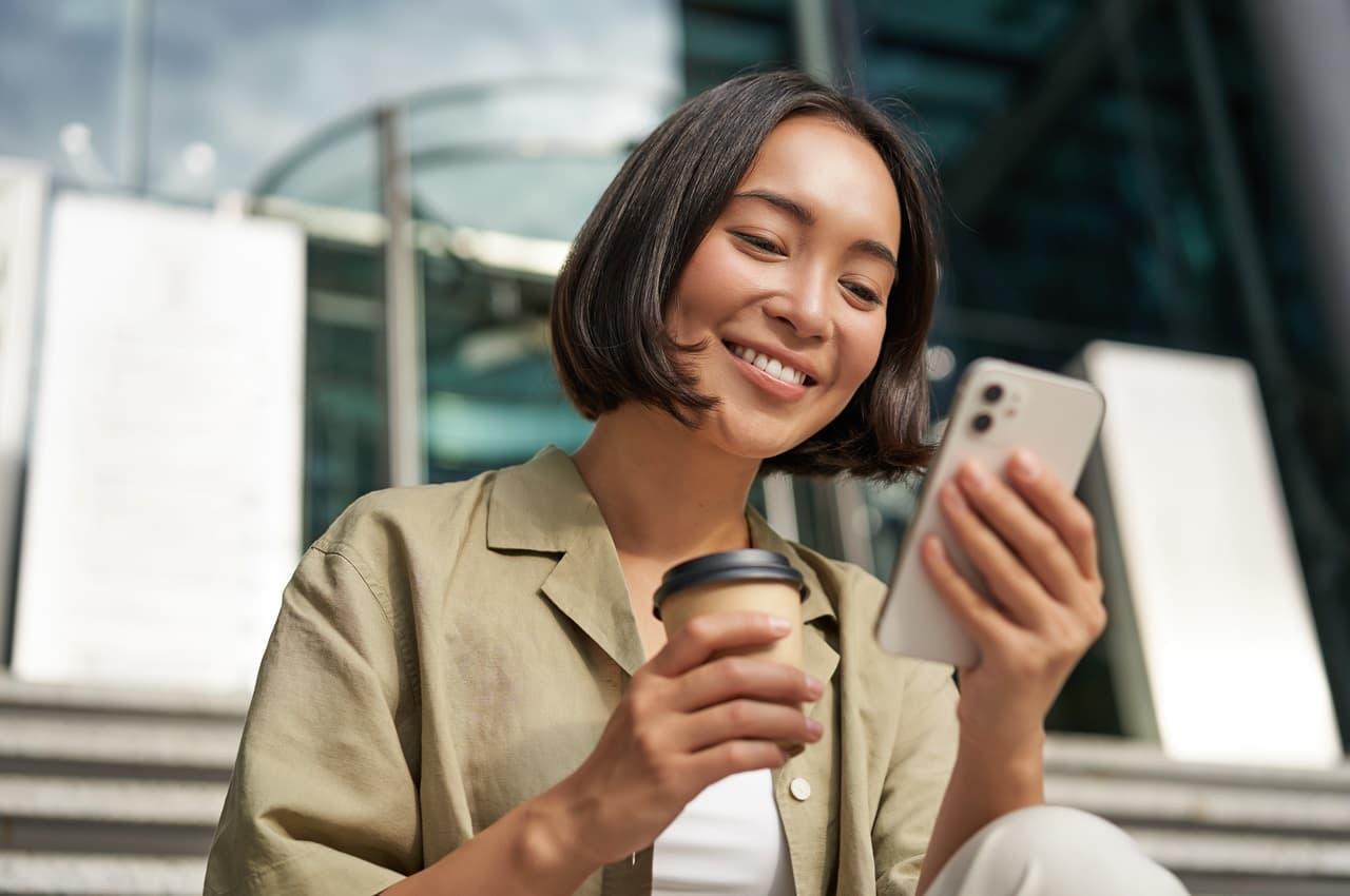 Mulher sorridente segurando um copo de café em uma mão e o celular em outra. Ela está lendo sobre averbação de empréstimo consignado.