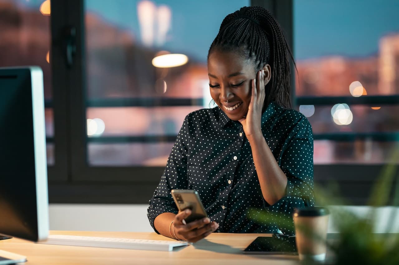 Mulher sorridente digitando no celular e apoiando uma das mãos na bochecha. Ela veste blusa de bolinhas preta e branca e está no escritório do trabalho.