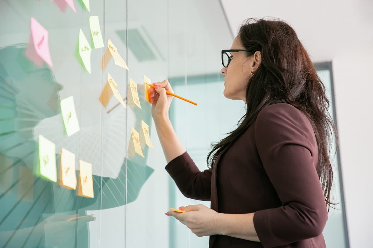 Mulher com expressão concentrada preenchendo post-it em um quadro transparente para um planejamento de vida financeira.