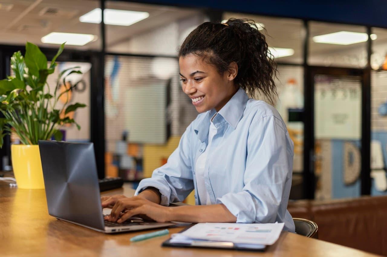 Mulher sorridente veste camisa social azul e digita no laptop em cima da mesa do escritório e ao lado de uma prancheta de reajuste salarial.