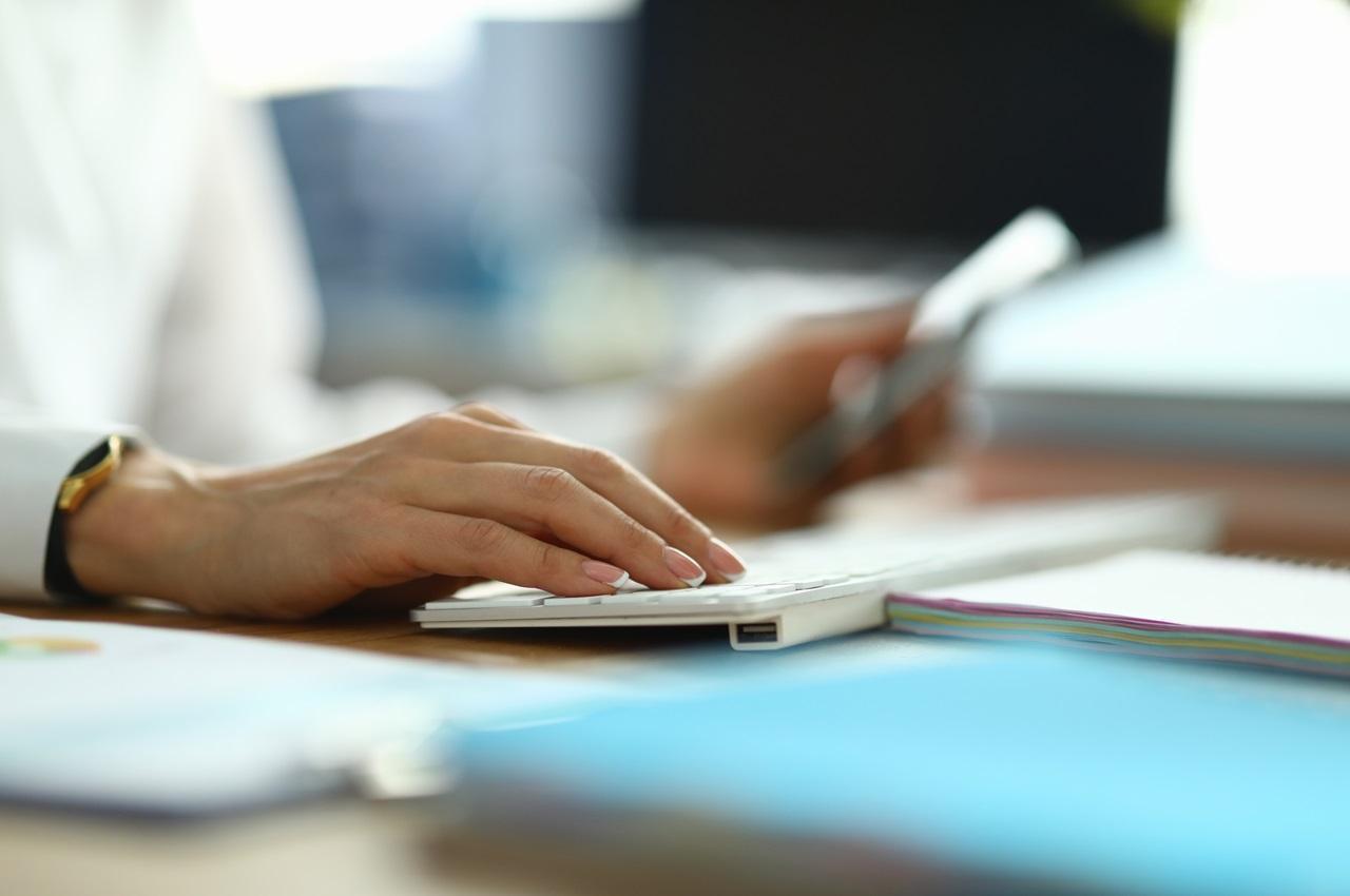 Foco em mão feminina digitando em teclado branco de computador. Ao lado de teclado tem pastas na cor azul e a pessoa utiliza uma calculadora.