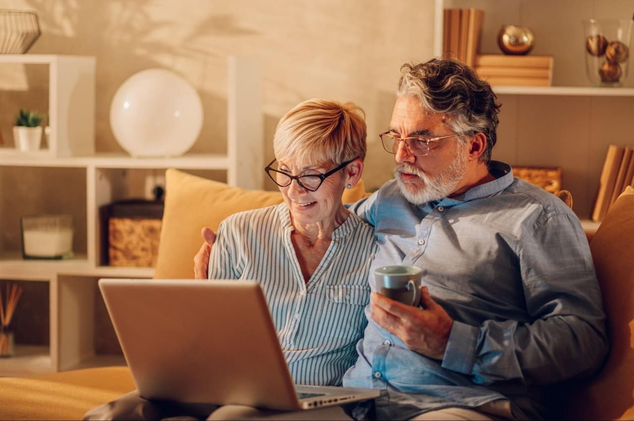 Casal sênior de uma mulher e um homem abraçados no sofá de casa, enquanto olham para a tela de um computador. O homem segura uma xícara de café nas mãos.