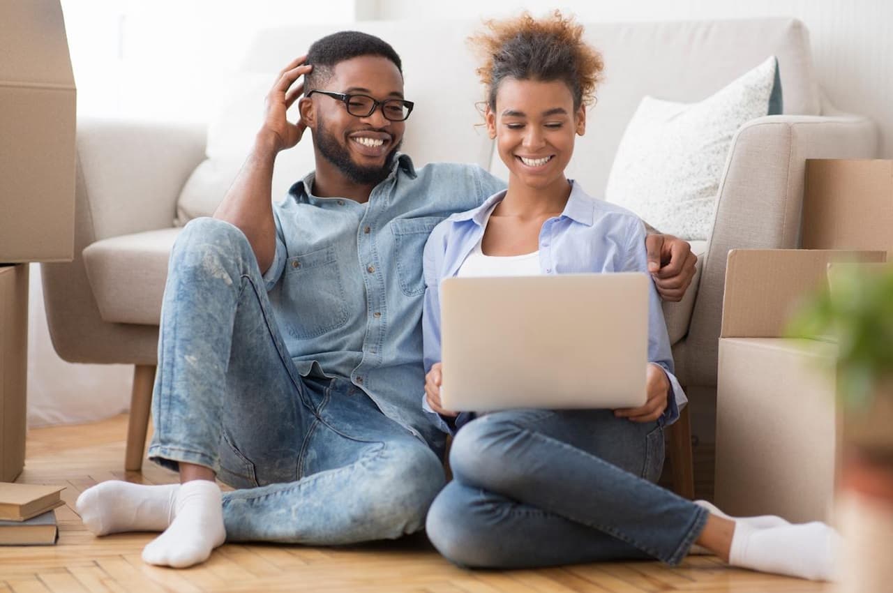 Casal (um homem e uma mulher) sorridente sentado no chão. Eles vestem calças jeans, meias brancas e camisas azuis básicas. A mulher segura um laptop em cima do colo