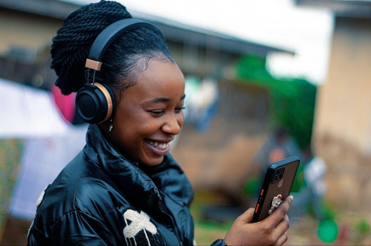 Mulher sorridente com fone de ouvido sem fio e procura cheque especial ou empréstimo pelo celular. A mulher veste jaqueta grande preta, brincos de prata e está em pé em uma calçada.