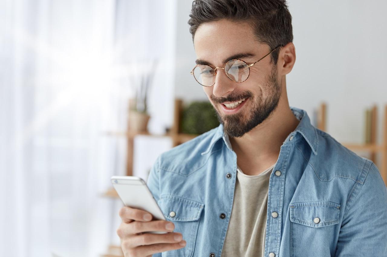 Homem sorridente utilizando celular para aumentar o score. O homem tem cabelos e barba castanhos curtos, veste camisa jeans e óculos de grau.