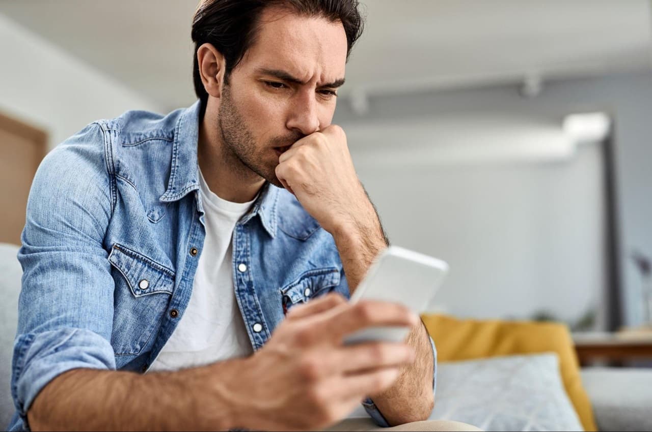 Homem com expressão preocupada, sentado no sofá e uma mão apoiada na boca, enquanto pesquisa como quitar dívidas no celular. 