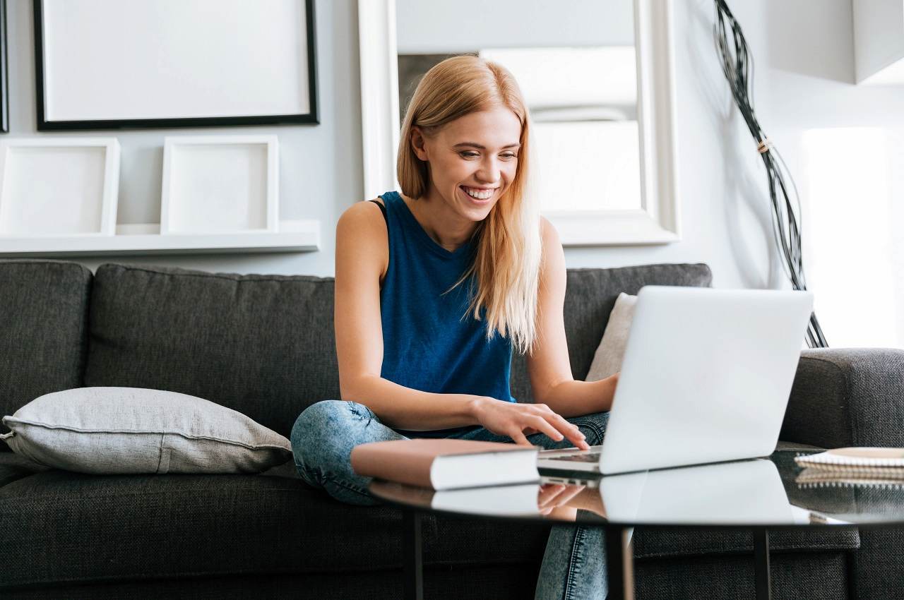 Mulher loira sorridente sentada no sofá, acessando contrato de empréstimo pessoal no computador. Ela veste regata azul e calça jeans.