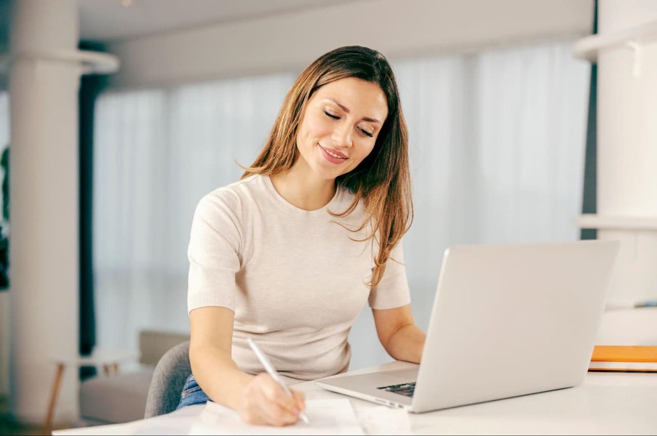 Mulher sorridente, vestindo camisa básica branca, calça jeans e sentada na sua mesa de escritório escrevendo e usando um computador.