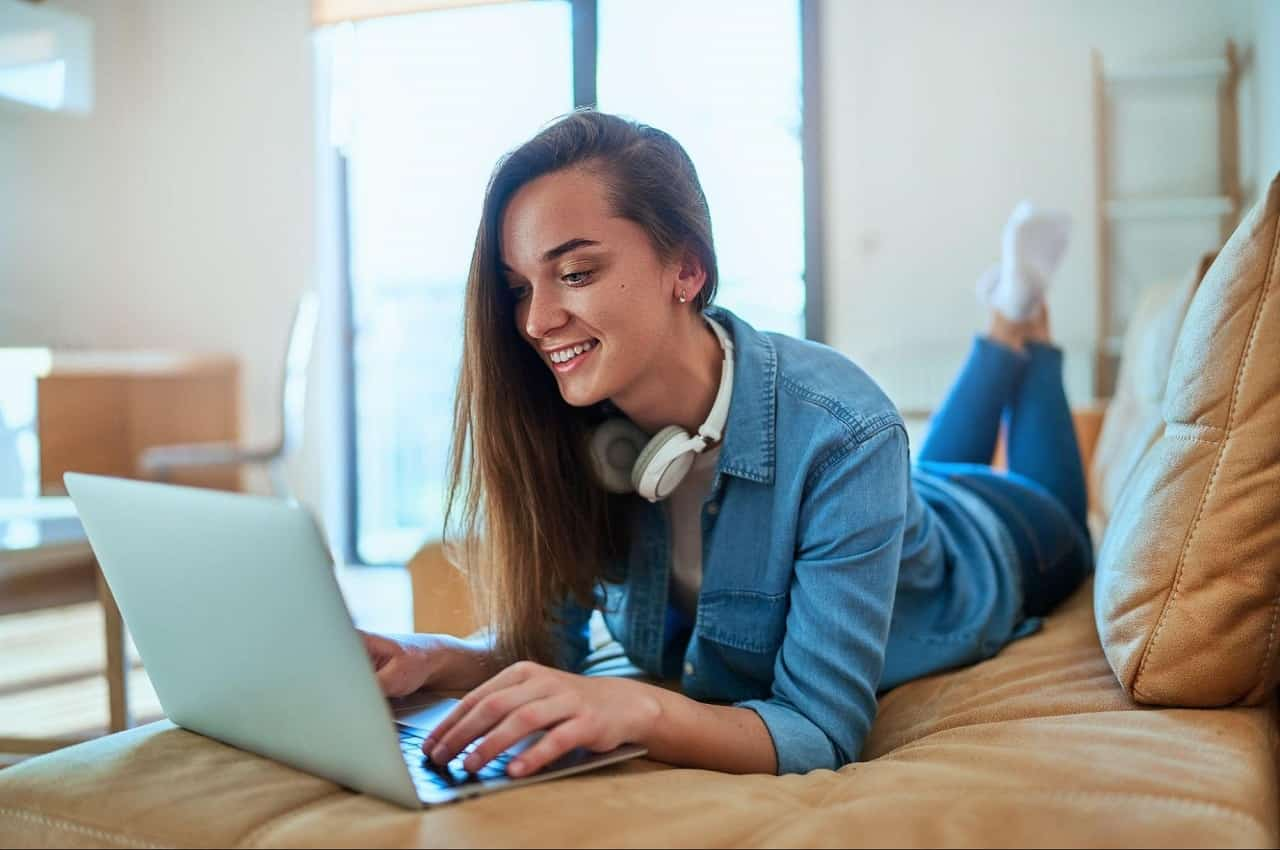 Mulher sorridente de cabelos castanhos longos e fones de ouvido bluetooth, pesquisando empréstimo pessoal online no laptop. Ela está deitada de barriga para baixo em um sofá, vestindo conjunto de roupas jeans.