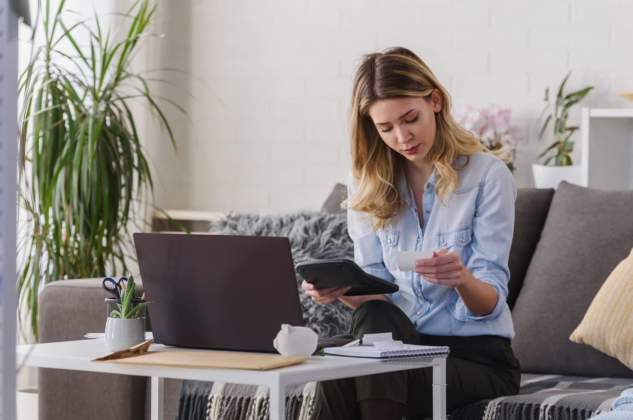 Mulher loira com expressão concentrada segurando calculando juros do empréstimo pessoal com calculadora e anotações. Ela está sentada no seu sofá, utilizando um computador e vestindo roupas casuais.