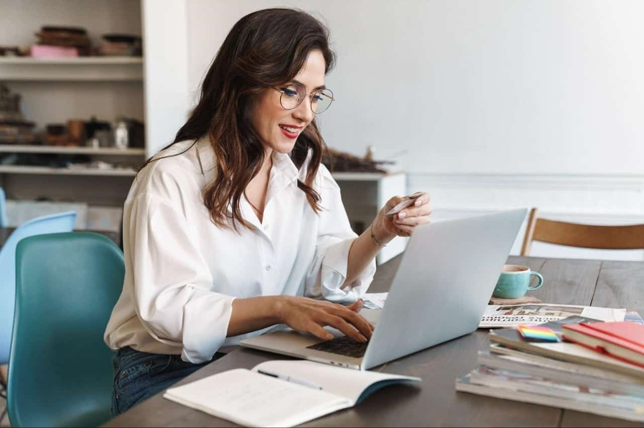 Mulher sorridente de cabelos castanhos soltos, blusa social branca e óculos de grau, utilizando computador para pedir empréstimo pessoal. 