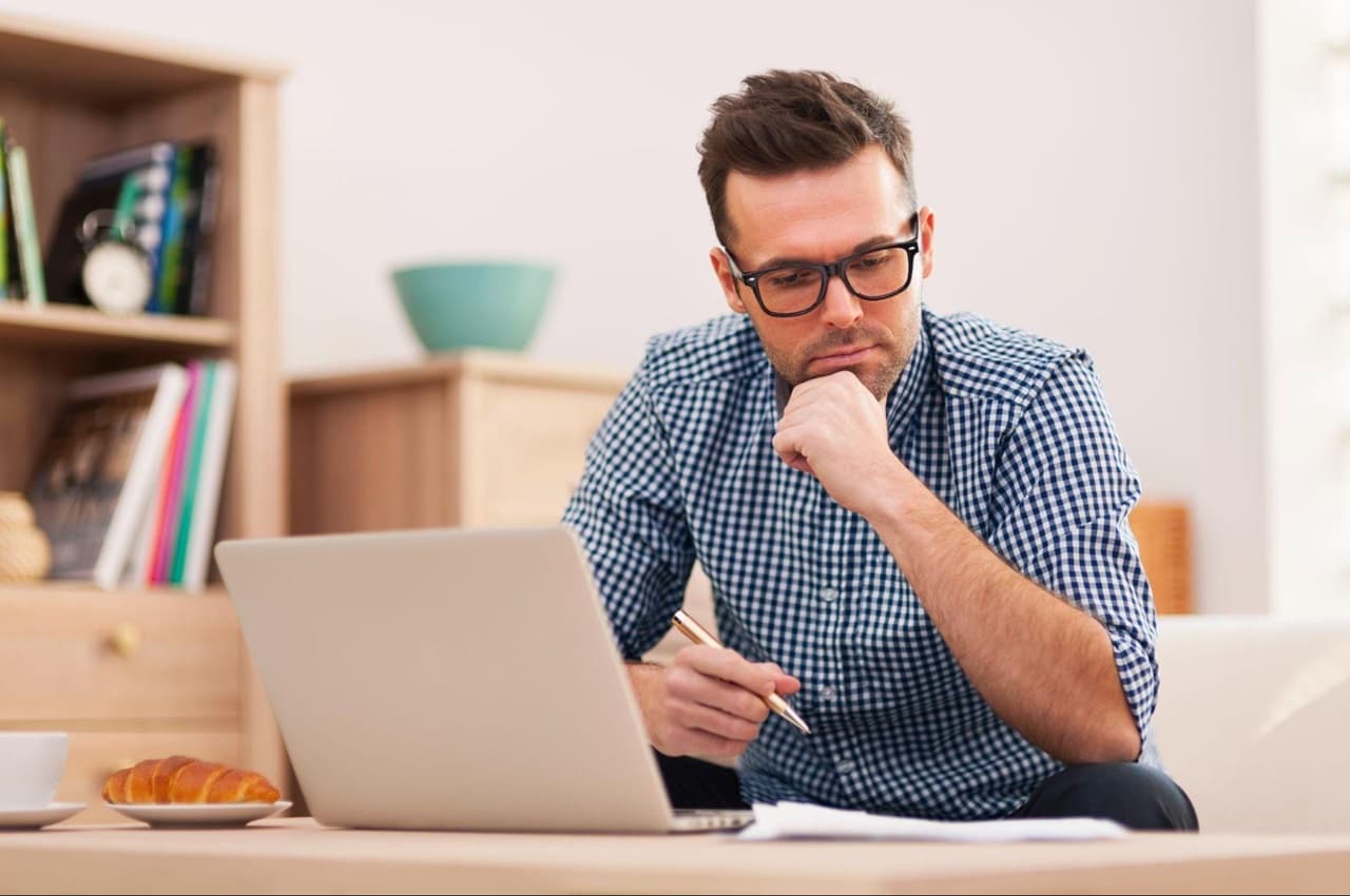 Homem com expressão concentrada, camiseta quadriculada azul e acessando portabilidade de empréstimo pelo computador. Ele está sentado no sofá e segura uma caneta.