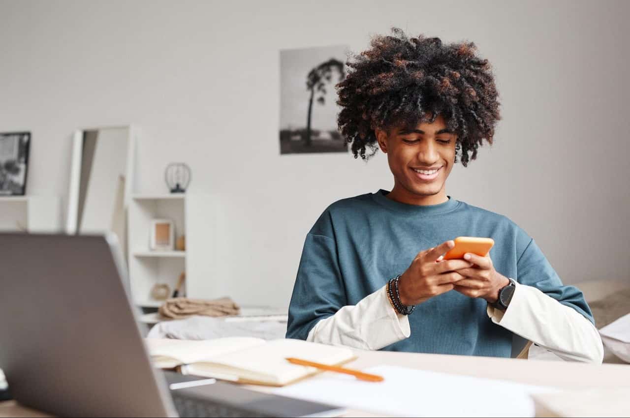 Homem sorridente, sentado em seu escritório utilizando celular e computador. Ele veste blusa de mangas brancas e, por cima, uma camisa básica azul.