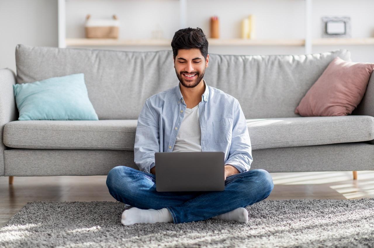 Homem sorridente sentado no chão vestindo calça jeans, meias e blusa social azul. Ele utiliza seu computador no colo.