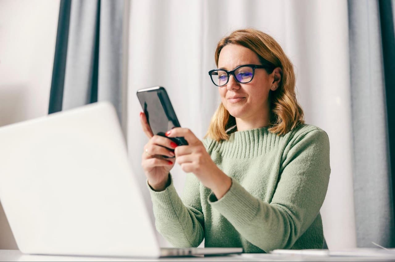 Mulher sorridente de cabelos loiros curtos, vestindo suéter gola alta verde e digitando no celular. Ela está sentada no seu escritório em frente ao computador.