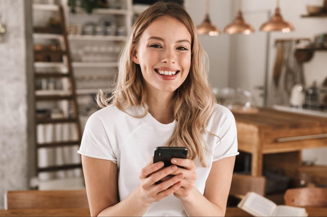 Mulher sorridente de cabelos loiros longos, vestindo blusa básica branca e segurando celular. Ao fundo, tem uma cozinha feita de madeira.