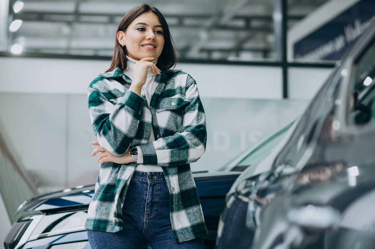 Mulher sorridente com as mãos apoiadas no queixo observando veículo para comprar direto com proprietário. Ela veste sobretudo xadrez e calça jeans.