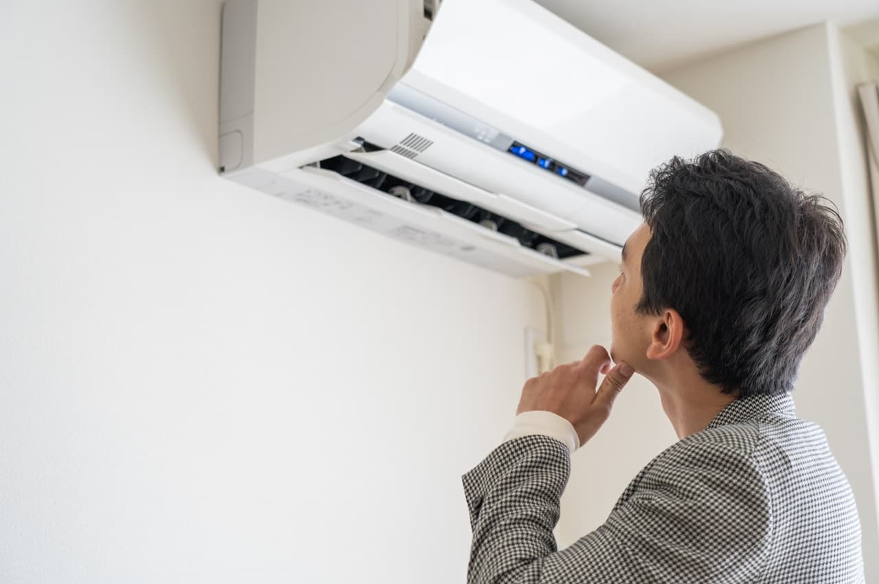 Homem de cabelos castanhos com a mão esquerda no queixo e expressão preocupada olhando para um ar-condicionado que consome energia.