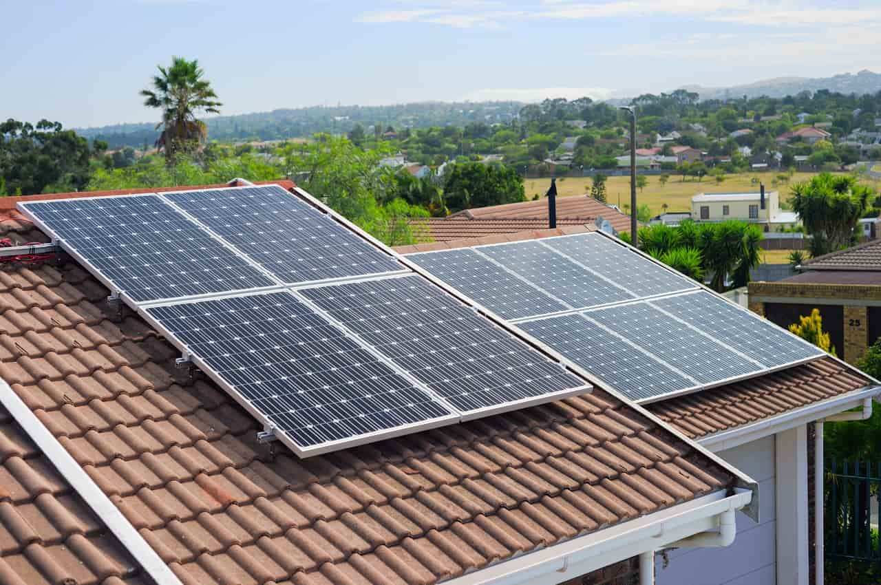 Telhado de uma residência de cor branca com placas solares instaladas. Ao fundo, tem um bairro com várias árvores e outras residências. 