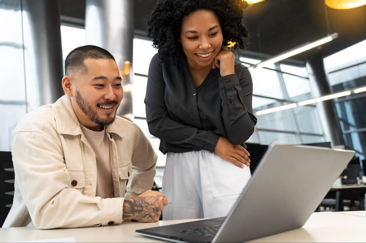 Homem e mulher sorridentes observam tela de laptop do trabalho no escritório da empresa. A mulher veste calça de alfaiataria branca e camisa social preta e o homem veste jaqueta jeans bege.