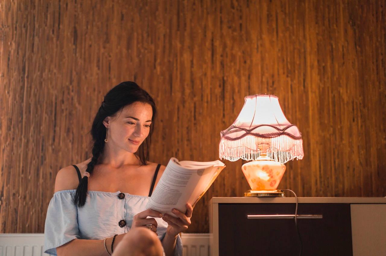 Mulher sorridente com blusa ombro a ombro azul e trança lateral. Ela lê um livro próxima ao abajur aceso pela energia elétrica.