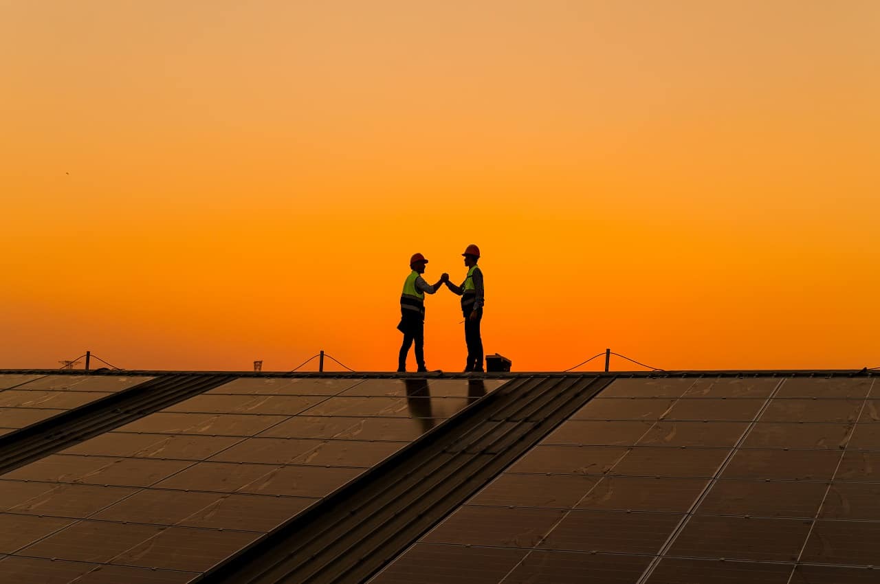 Duas pessoas dando um aperto de mão em cima de uma instalação de energia solar. Ao fundo tem o por de sol.