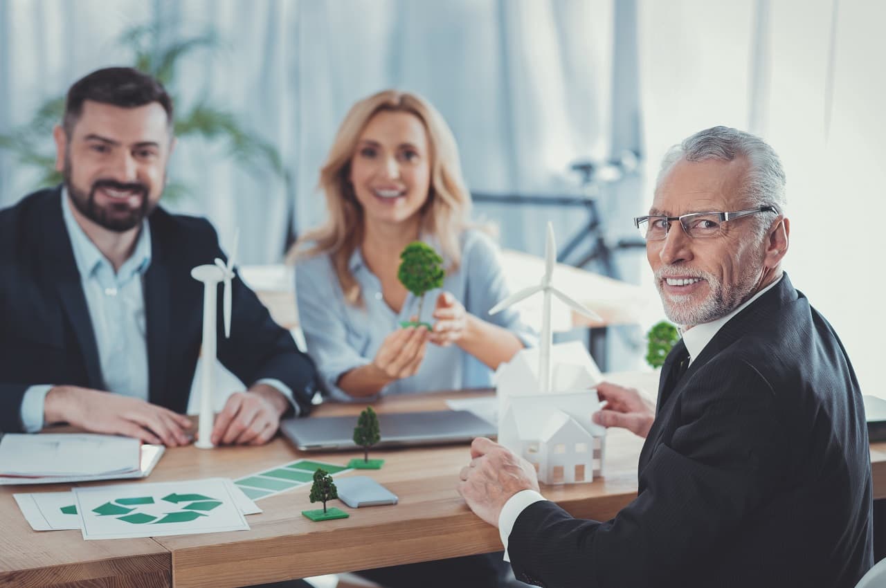 Dois homens e uma mulher sorridentes e com roupas formais, sentados em frente de uma mesa com papéis sobre empresa sustentável e árvores em miniatura. 