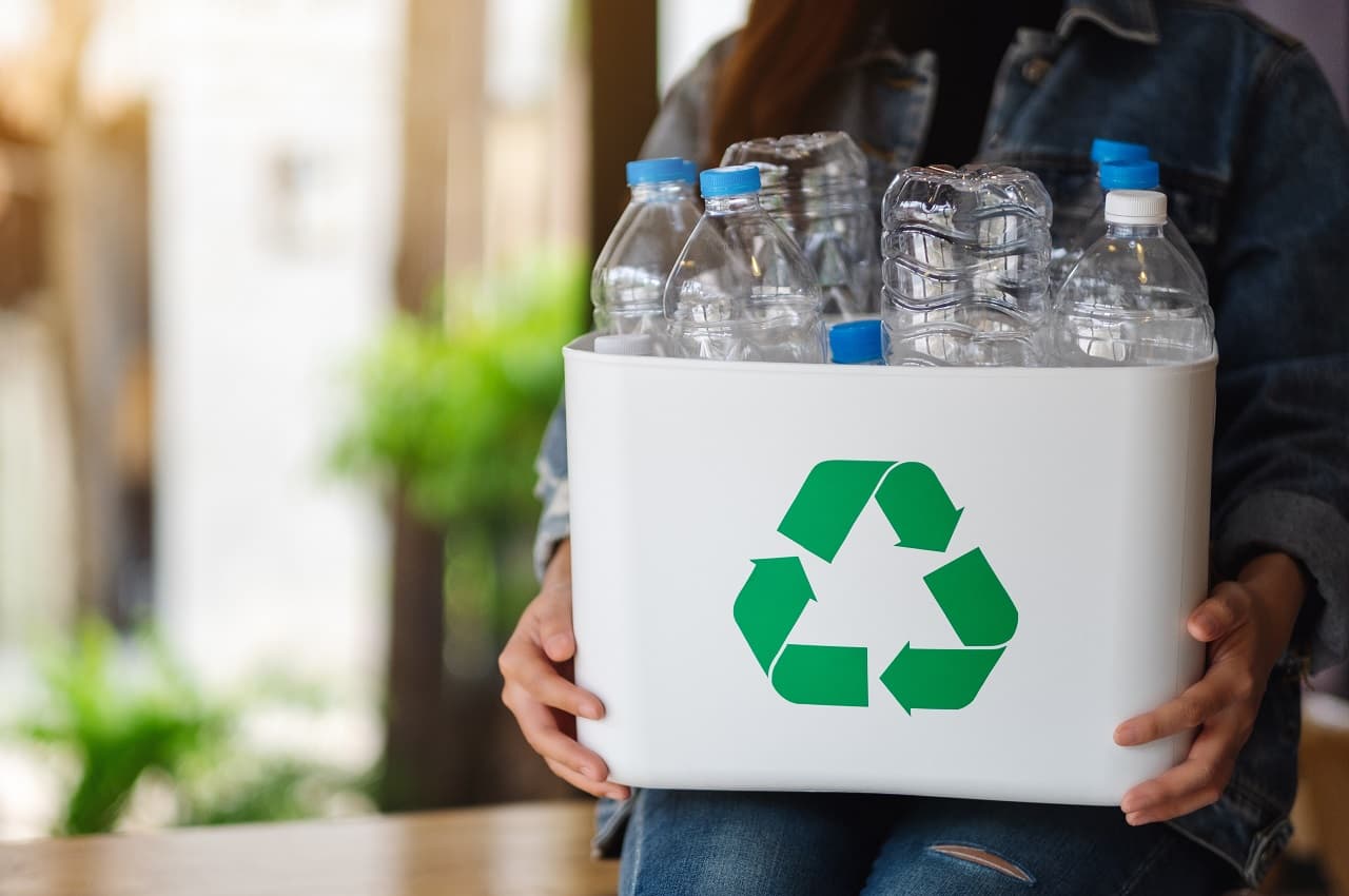 Pessoa segurando uma caixa com símbolo da reciclagem e nela contém garrafas PET vazias para reciclar.