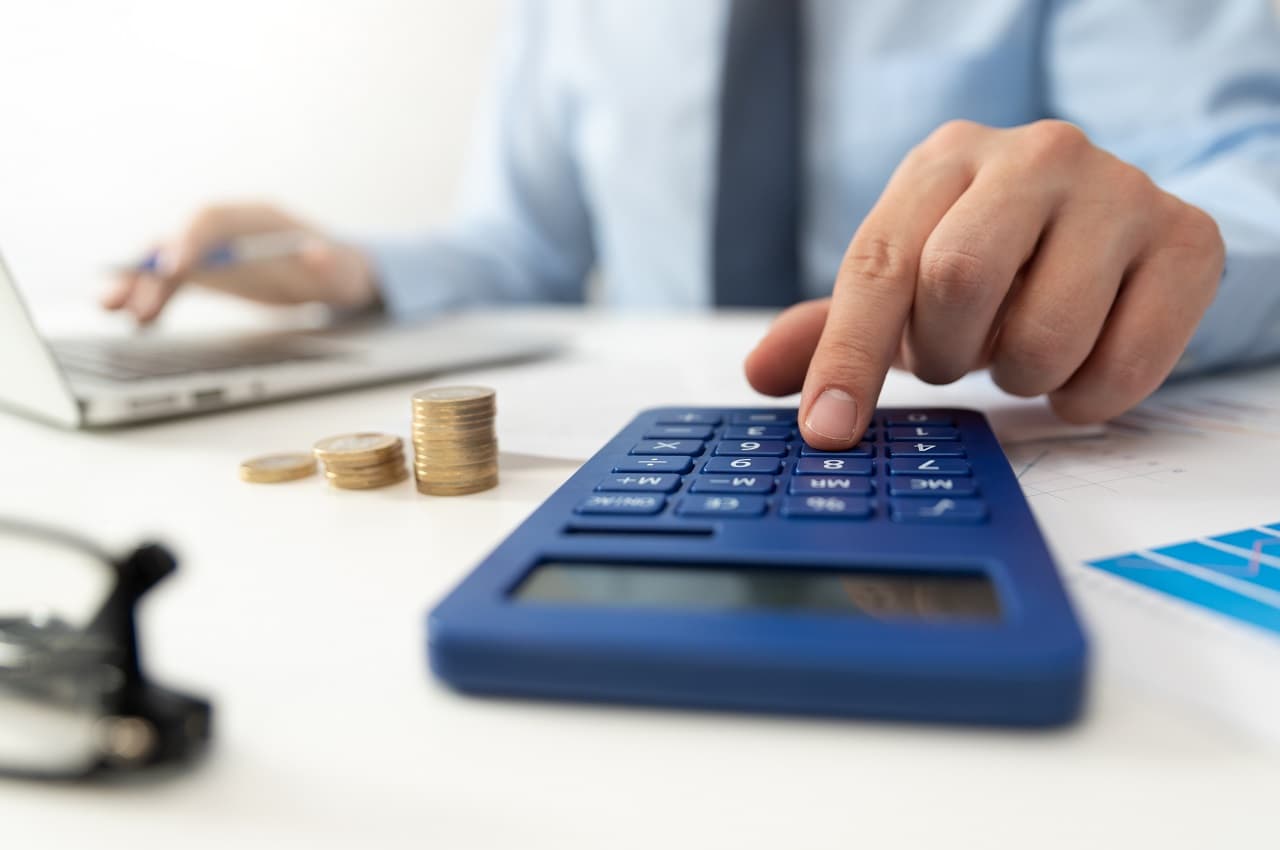 Pessoa clicando em calculadora e usando um computador que está na mesa. Ao lado do computador, tem uma pilha de moedas e contas de energia.