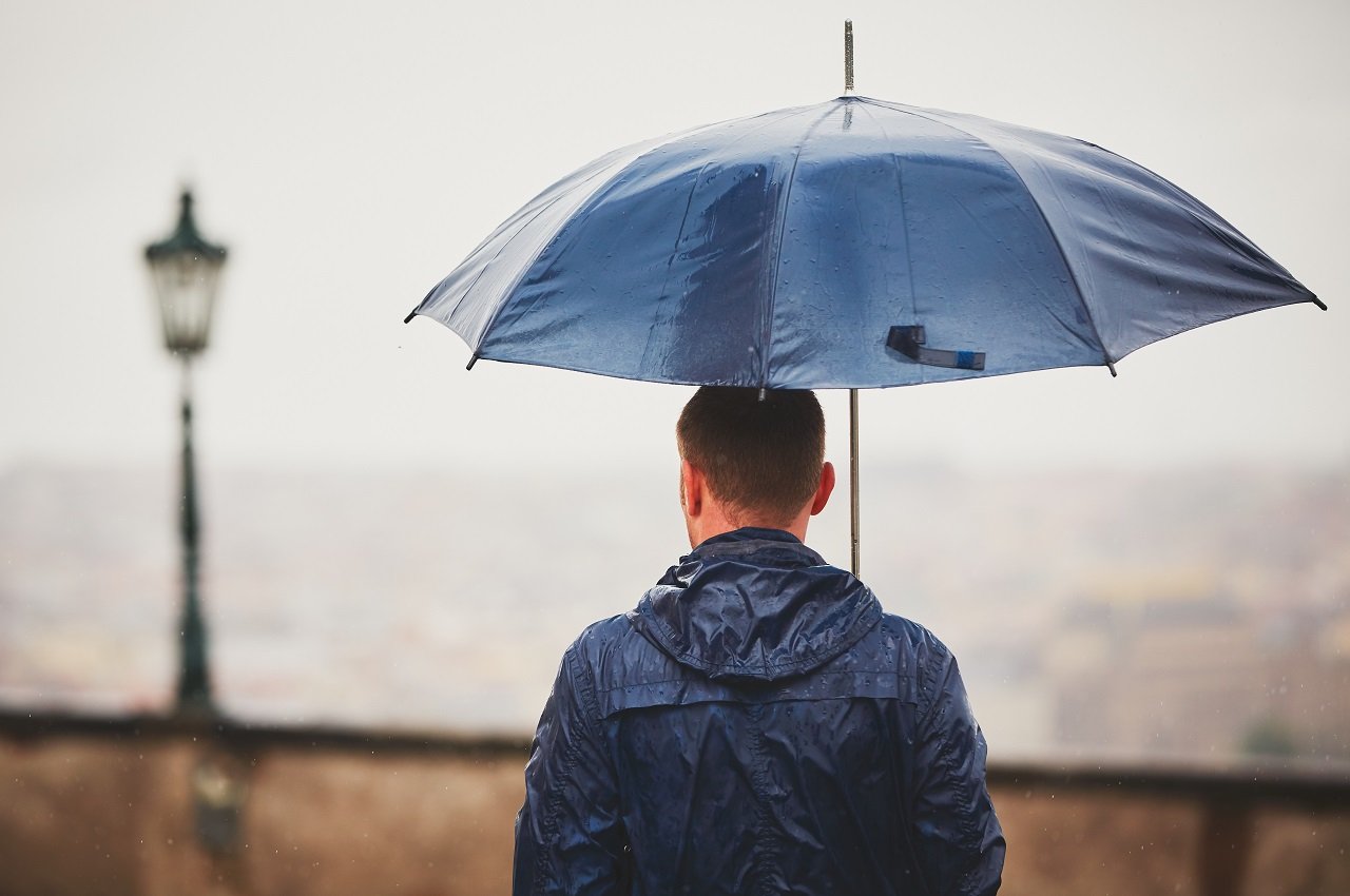 Homem de costas para a imagem e na chuva, vestindo uma jaqueta molhada e segurando um guarda-chuva azul escuro.