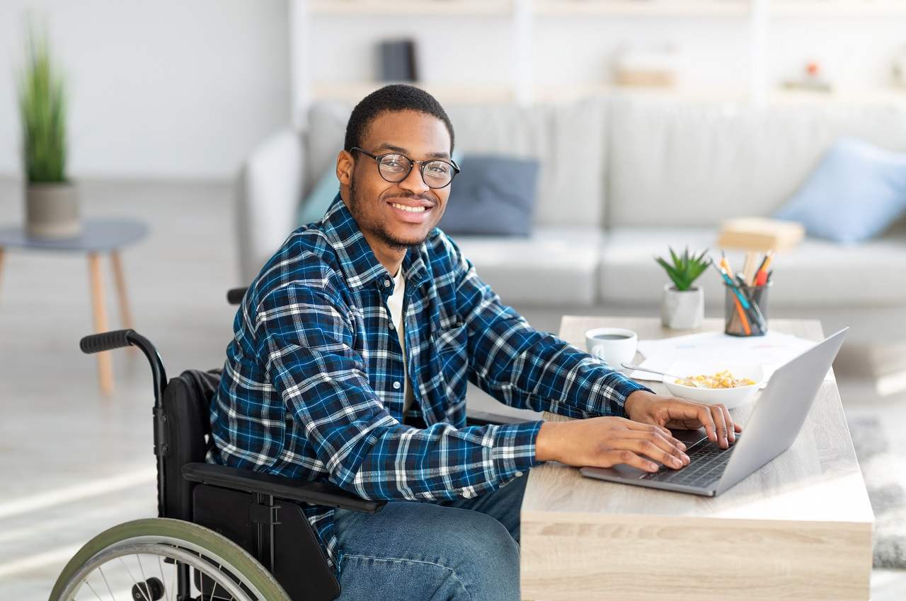 Homem sorridente e cadeirante, vestindo blusa xadrez azul e digitando em um computador na sua casa.