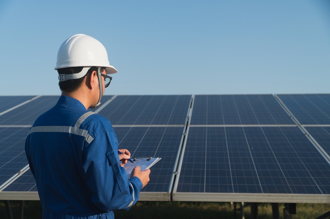 Homem de costas com capacete branco e roupa de segurança azul olhando para placas solares instaladas e anotando na prancheta de trabalho.