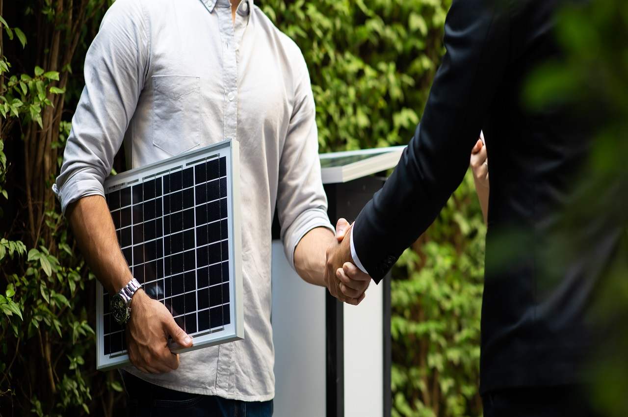 Aperto de mão entre dois homens (seus rostos não aparecem na imagem). Uma das pessoas está segurando um painel de energia solar.