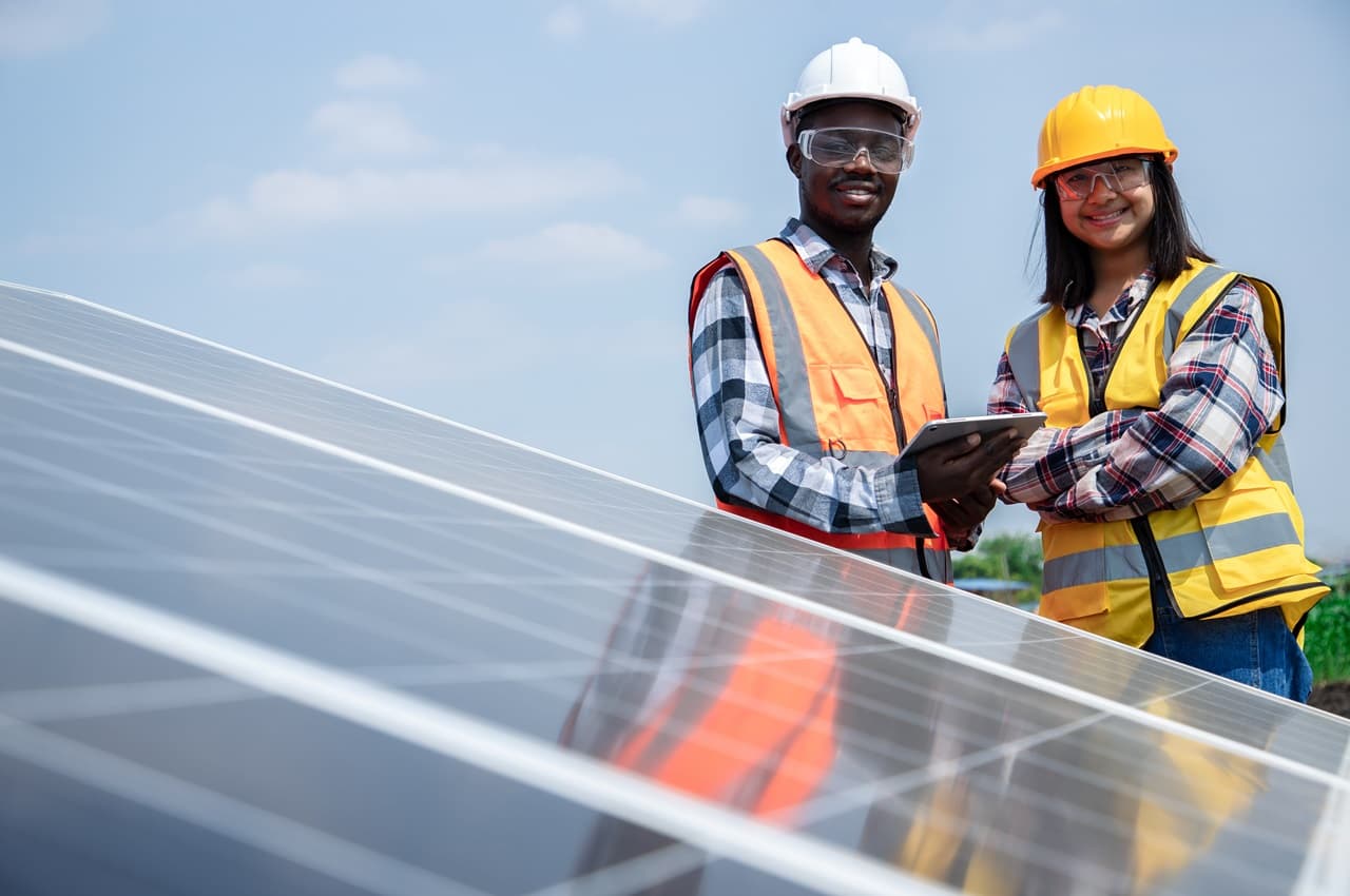 Homem e mulher sorridentes e paramentados com EPIs (capacete, óculos e colete de segurança) em frente a painéis solares.