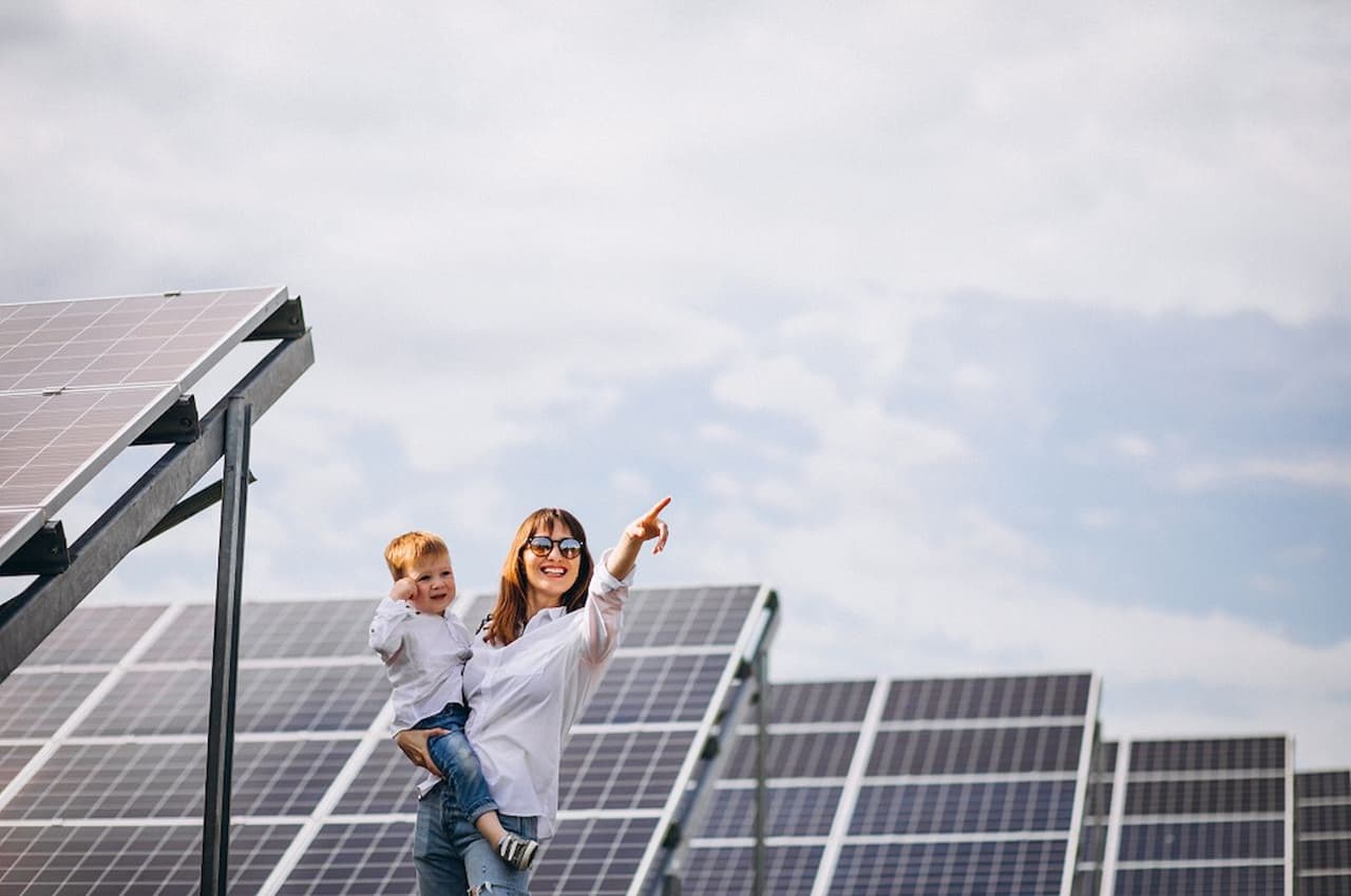 Mulher de cabelos castanhos e óculos de sol, segurando um bebê no colo e apontando para frente. Ao fundo tem várias placas solares instaladas.