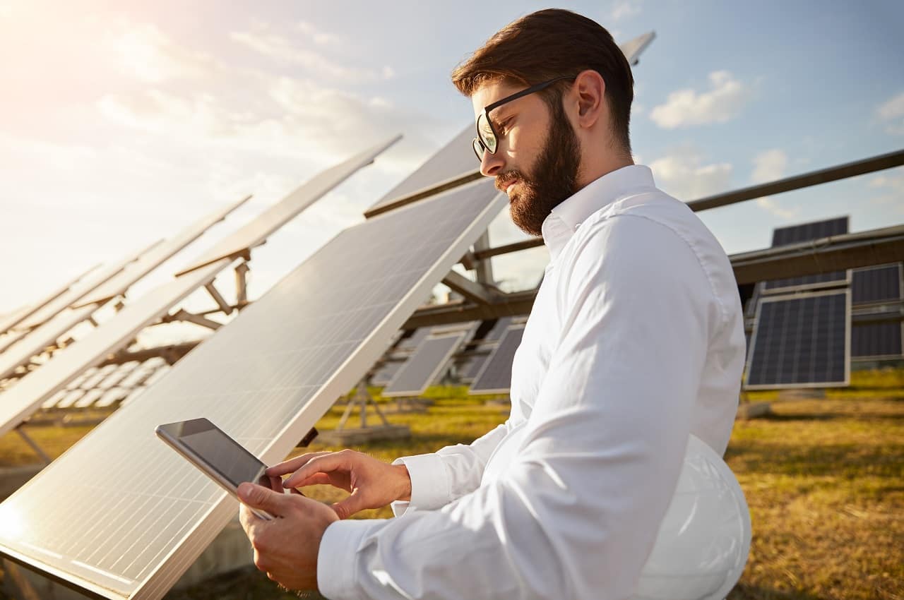 Homem com expressão concentrada segurando um capacete de proteção e digitando no celular. Ele está próximo a painéis solares instalados.