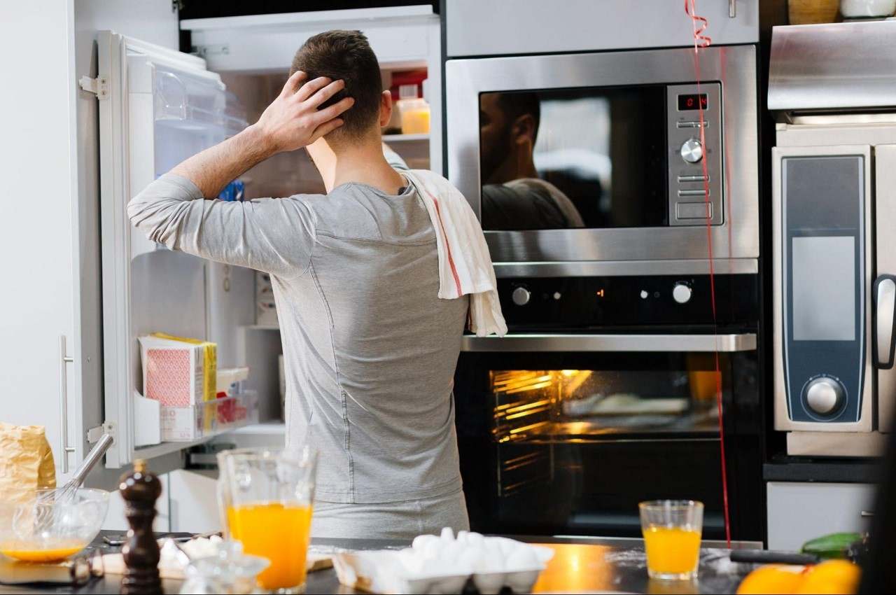 Homem abrindo a geladeira e de costas para a bancada de uma cozinha que tem um suco e uma bandeja de ovos. 